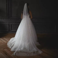 Load image into Gallery viewer, A bride wearing a long, white wedding dress with a sparkly veil stands in a dimly lit room, facing away from the camera.