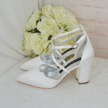 Load image into Gallery viewer, A pair of white strappy high-heeled shoes with a metallic strap design, placed next to a bouquet of white roses against a pink brick wall.