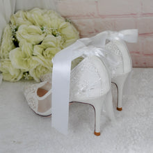 Load image into Gallery viewer, A pair of white lace high-heeled shoes with a white ribbon tied around the toe, placed on a white surface next to a bouquet of white flowers.