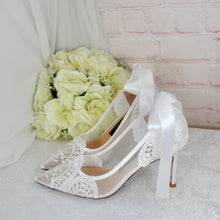 Load image into Gallery viewer, A pair of white lace high-heeled shoes with a ribbon tied around the toe, placed on a white surface next to a bouquet of white flowers.