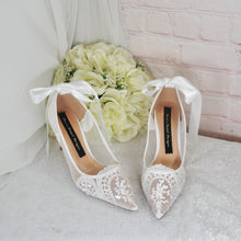 Load image into Gallery viewer, A pair of white lace wedding shoes with bows, placed next to a bouquet of white roses, against a white brick wall.
