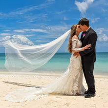 Load image into Gallery viewer, A bride and groom embrace on a beach, with the bride's veil flowing behind them.