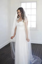 Load image into Gallery viewer, A woman in a white wedding dress stands in a room with a window, her veil flowing behind her.