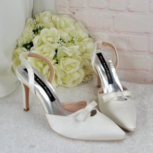 Load image into Gallery viewer, A pair of white high-heeled shoes with bows on the toe, placed next to a bouquet of white roses on a white surface.