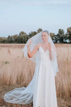 Load image into Gallery viewer, A woman in a white wedding dress stands in a field, her veil blowing in the wind.
