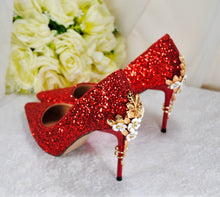 Load image into Gallery viewer, A pair of red glitter high-heeled shoes with gold floral embellishments, placed on a white surface with a bouquet of white roses in the background.