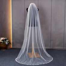 Load image into Gallery viewer, A white veil with a long, flowing train is displayed on a mannequin, with a vase of flowers visible to the left.