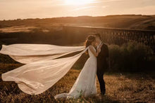 Load image into Gallery viewer, A couple embracing in a field at sunset, with the bride's flowing white dress and veil creating a romantic atmosphere.