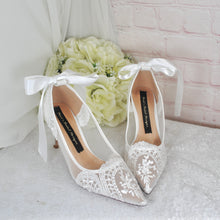 Load image into Gallery viewer, A pair of white lace wedding shoes with bows, placed on a white surface next to a bouquet of white flowers.