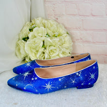 Load image into Gallery viewer, A pair of blue shoes with white stars on them, placed next to a bouquet of white flowers against a pink brick wall.