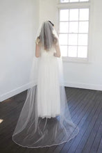 Load image into Gallery viewer, A woman in a white wedding dress stands in a room with a large window, her back to the camera.
