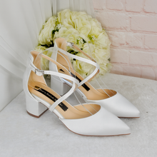 Load image into Gallery viewer, A pair of white strappy heeled shoes with a strap across the ankle, placed on a white surface next to a bouquet of white flowers.