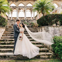 Load image into Gallery viewer, A bride and groom stand on stone steps in a lush garden, with the bride's veil flowing behind them.