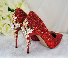 Load image into Gallery viewer, A pair of red glitter high-heeled shoes with gold floral embellishments, placed on a white surface with a bouquet of white roses in the background.