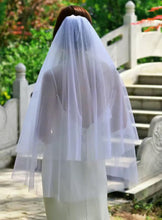Load image into Gallery viewer, A woman wearing a white wedding veil is walking on a stone path in a garden setting.
