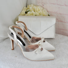 Load image into Gallery viewer, A pair of white high-heeled shoes with bows on the toe, placed next to a white clutch bag and a bouquet of white flowers.
