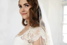 Load image into Gallery viewer, A woman in a white lace wedding dress with a veil stands in a room with a white background.
