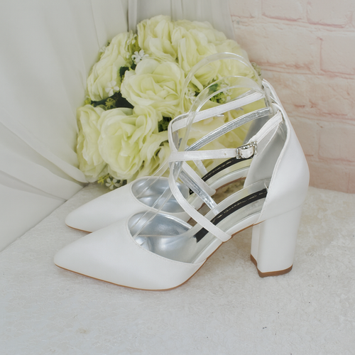 A pair of white strappy high-heeled shoes with a metallic strap design, placed next to a bouquet of white roses against a pink brick wall.