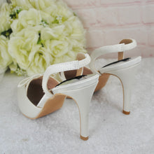 Load image into Gallery viewer, A pair of white high-heeled shoes with a bow detail, placed on a white surface next to a bouquet of white roses.
