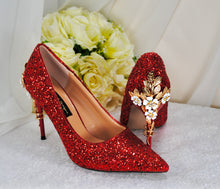 Load image into Gallery viewer, A pair of red glitter high-heeled shoes with gold embellishments, placed next to a bouquet of white roses.
