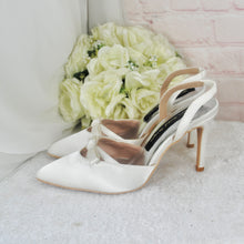 Load image into Gallery viewer, A pair of white high-heeled shoes with a bow detail, placed next to a bouquet of white roses on a white surface.
