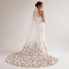 Load image into Gallery viewer, A woman in a white wedding dress with a long, flowing veil adorned with floral embroidery stands against a plain white background.
