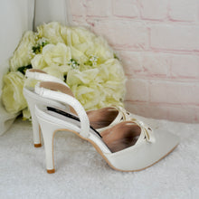 Load image into Gallery viewer, A pair of white high-heeled shoes with bows on the toe rests on a white surface, with a bouquet of white flowers in the background.
