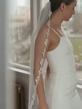 Load image into Gallery viewer, A woman in a white dress stands in front of a window, her hair adorned with a long, white veil decorated with delicate white leaves.

