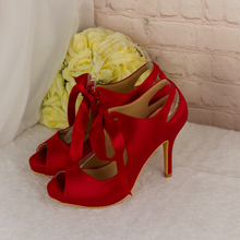 Load image into Gallery viewer, A pair of red high-heeled shoes with a bow on the toe, placed on a white surface next to a bouquet of yellow flowers.
