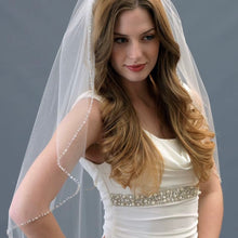 Load image into Gallery viewer, A woman with long, wavy brown hair wearing a white wedding dress with a beaded veil stands against a gray background.
