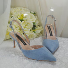 Load image into Gallery viewer, A pair of blue suede high-heeled shoes with a floral arrangement in the background.
