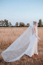 Load image into Gallery viewer, A bride in a white wedding dress stands in a field, her veil flowing behind her as she gazes into the distance.
