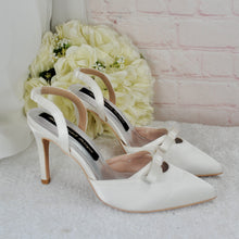 Load image into Gallery viewer, A pair of white high-heeled shoes with bows on the toe, placed next to a bouquet of white flowers against a pink brick wall.
