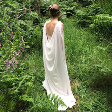 Load image into Gallery viewer, A woman in a long white dress stands in a lush, green forest, surrounded by tall grass and vibrant purple flowers.
