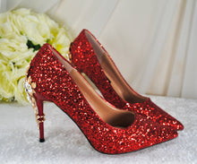 Load image into Gallery viewer, A pair of red glitter high-heeled shoes with gold embellishments, placed on a white surface next to a bouquet of white roses.
