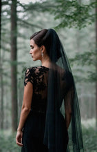 Load image into Gallery viewer, A woman wearing a black lace dress with a long black veil stands in a forest, facing away from the camera.
