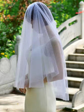 Load image into Gallery viewer, A bride wearing a white veil stands in front of a stone staircase in a garden setting.

