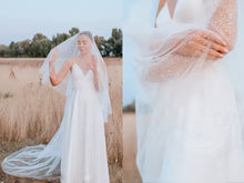 Load image into Gallery viewer, A bride in a white dress stands in a field, her veil blowing in the wind.
