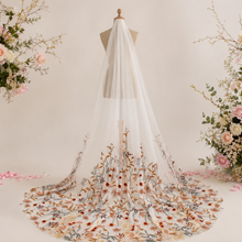 Load image into Gallery viewer, A white wedding veil with a floral pattern on the back, displayed on a mannequin head. The veil has a long, flowing train and a sheer, transparent fabric. The background features pink and white flowers, creating a romantic and elegant setting.
