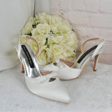 Load image into Gallery viewer, A pair of white high-heeled shoes with bows on the toe and heel, placed next to a bouquet of white roses on a white surface.
